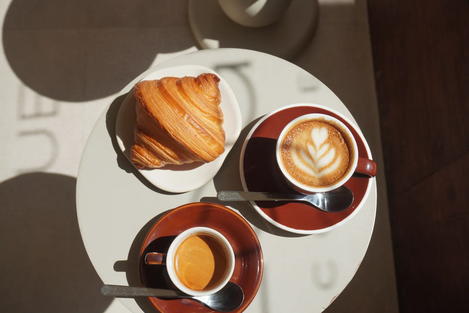 Overhead at Dialogues Coffee: a croissant, a cappuccino with leaf latte art in a red cup, and an espresso, in raking light.