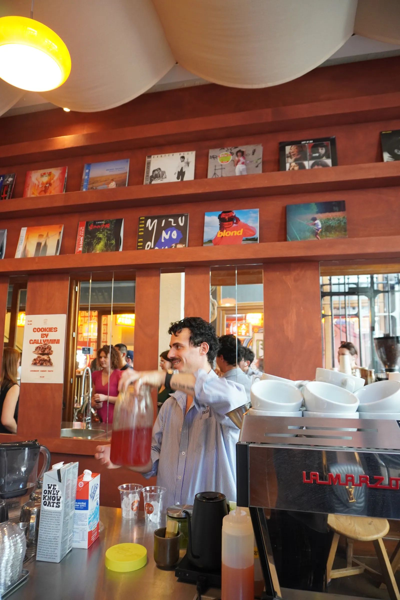 Behind the bar at Nami: vinyl sleeves on the shelf, the barista mid-pour under a yellow lamp.